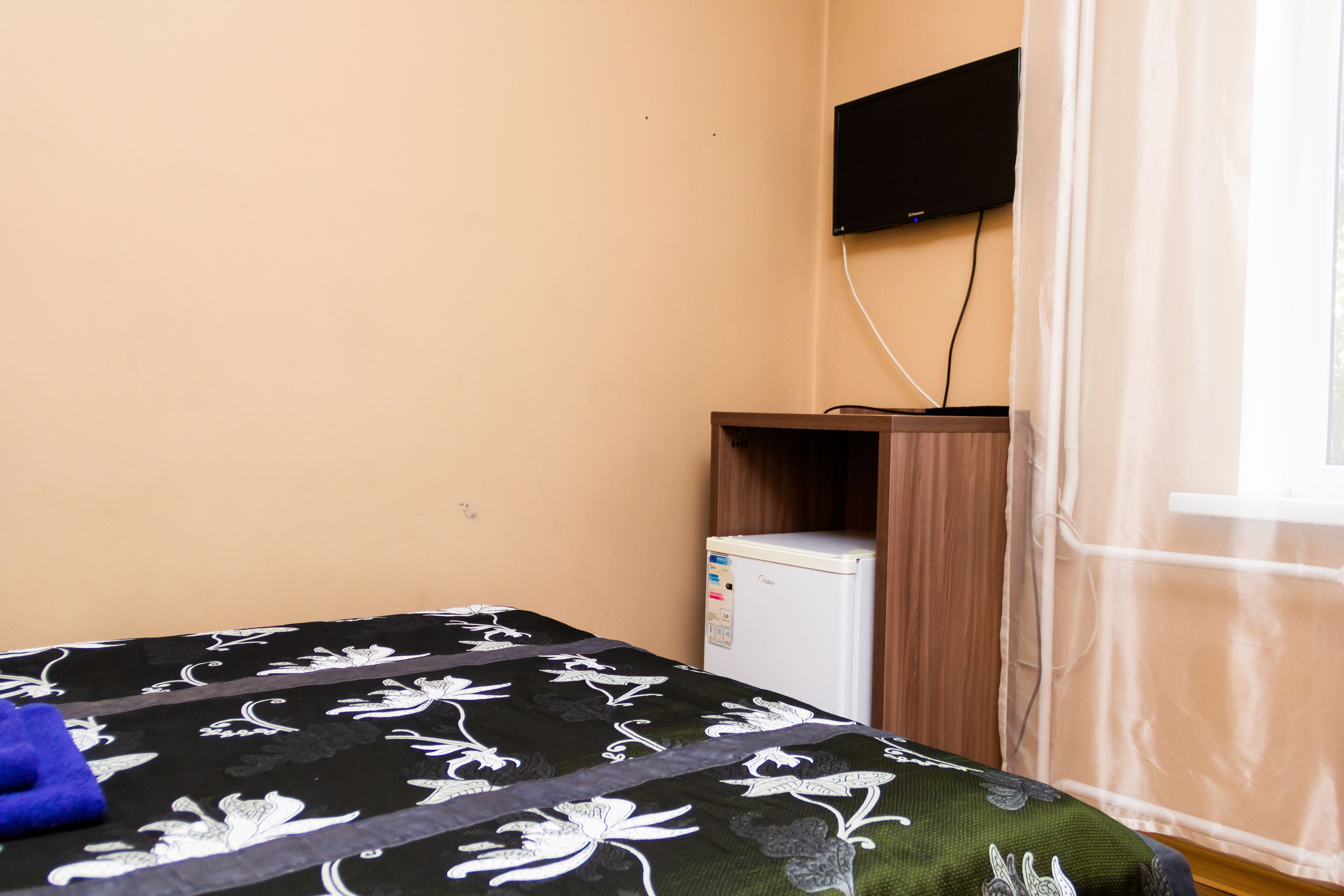 A room with a bed, a small refrigerator, a television mounted on the wall, and a window with curtains.