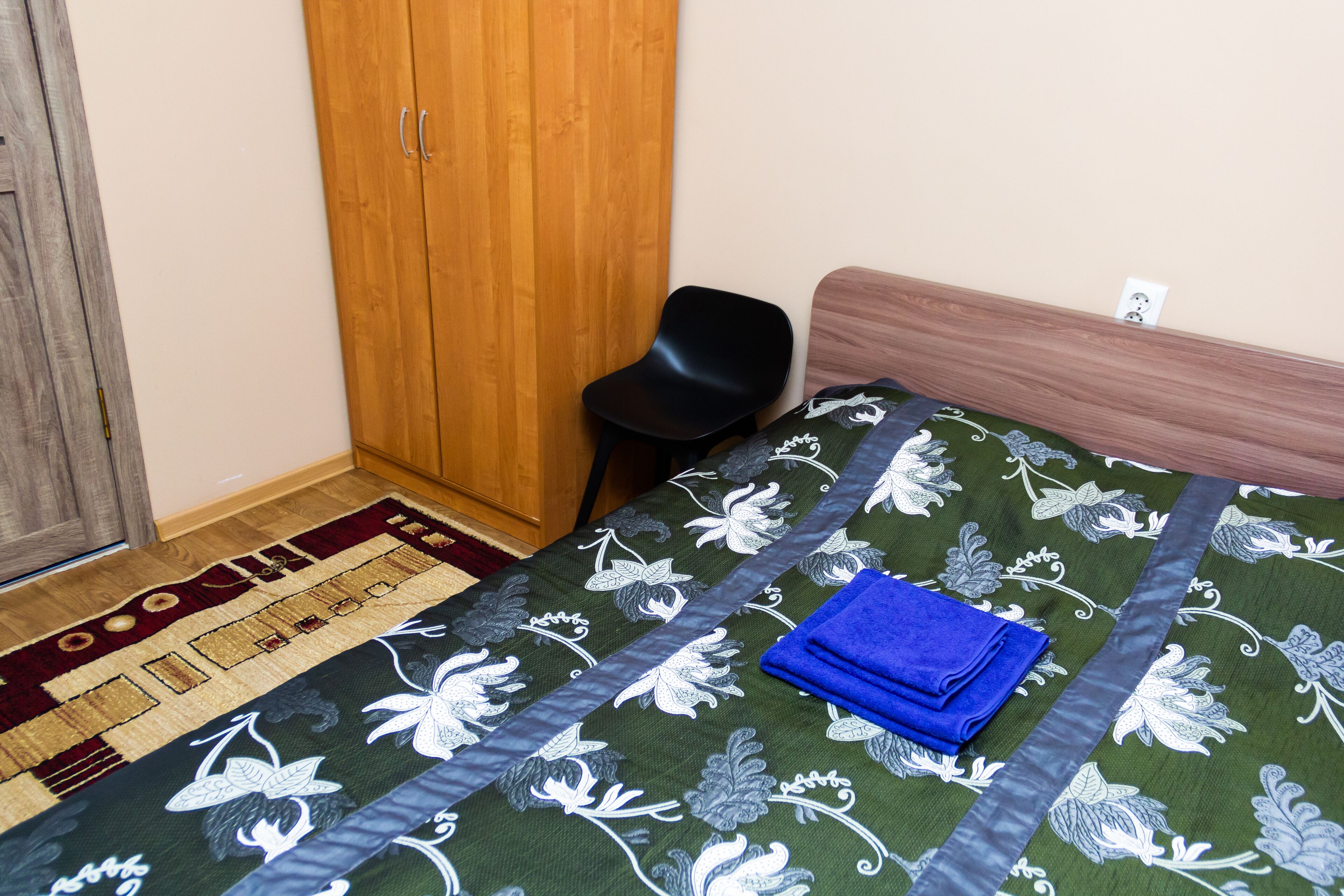 A bedroom with a bed, a wooden wardrobe, a black chair, and a patterned rug.