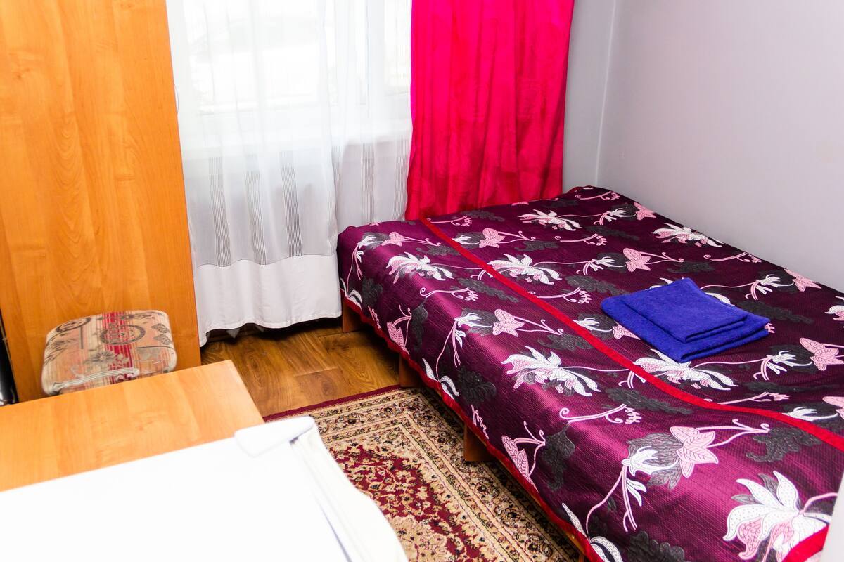 Una habitación pequeña con una cama, una mesa de madera, un tapete estampado y una ventana con cortinas traslúcidas y una cortina roja.