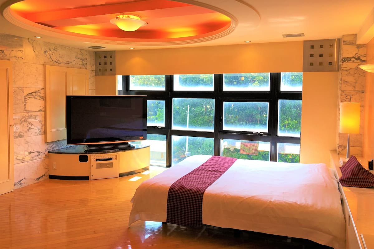 A hotel room with a large bed, a flat-screen TV, a marble wall, and a window with a view of greenery.