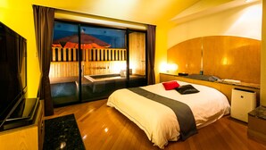A hotel room with a large bed, a TV, a small desk, and a view of a building with a red roof through a glass door.