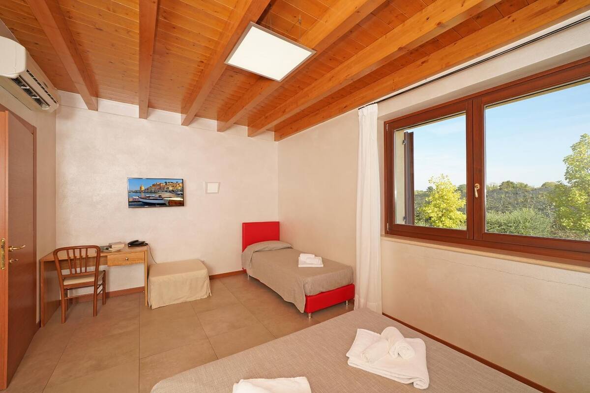 A bedroom with a wooden ceiling, a desk with a chair, a bed with red headboard, and a window with a view of greenery.