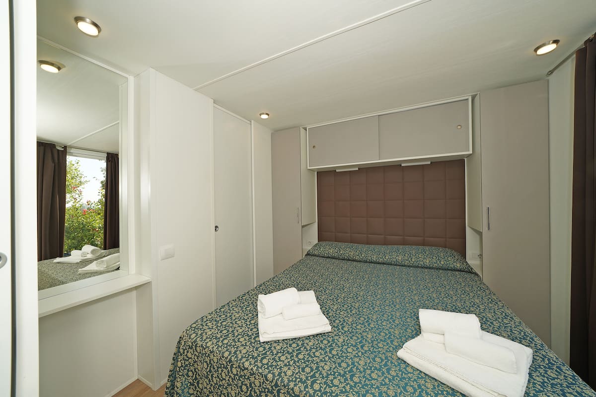 A hotel room with a bed, a window with curtains, a mirror, and a built-in headboard.