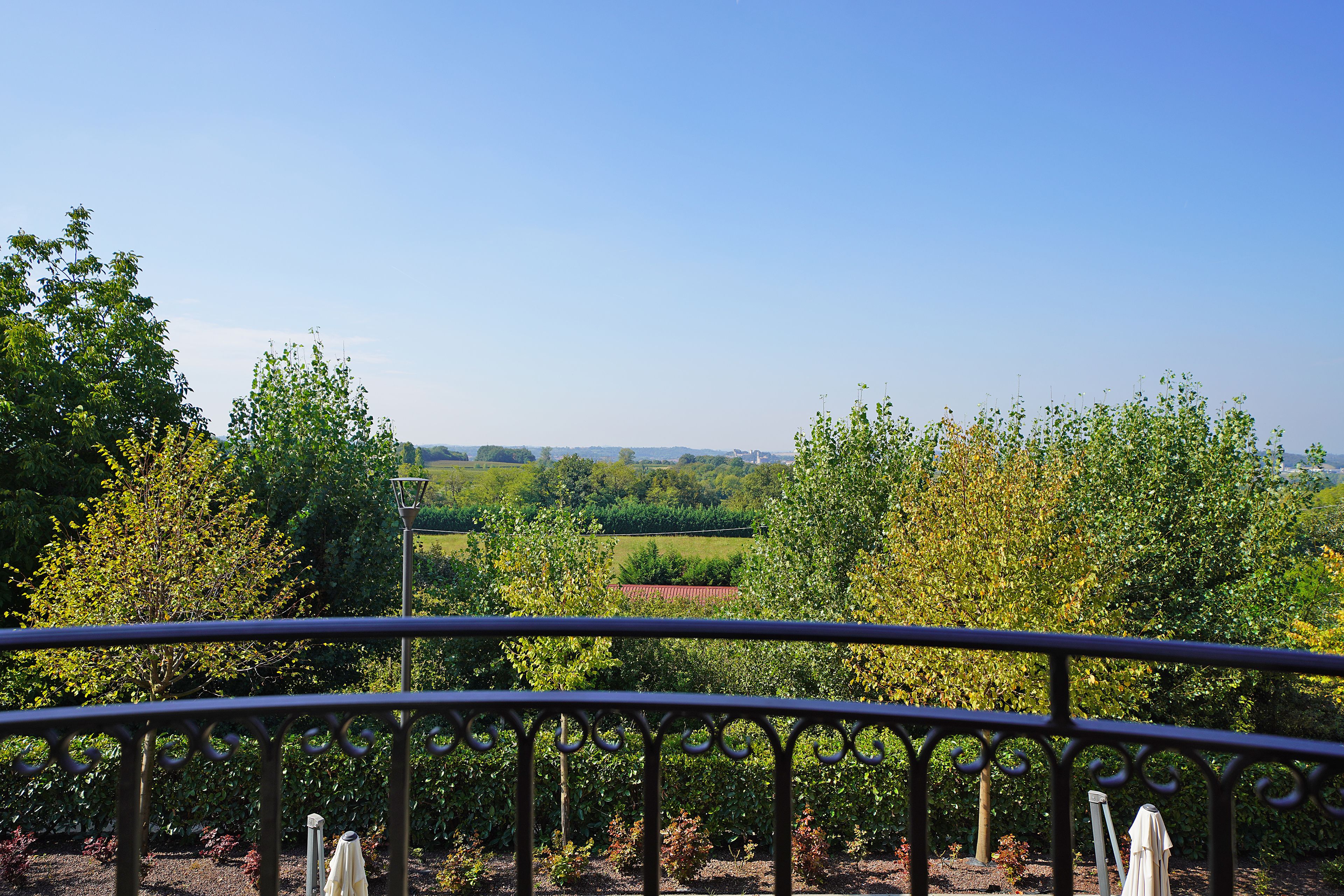 A balcony with a metal railing overlooking a garden with trees and a distant red-roofed building.