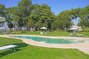 Piscina com borda curva, rodeada por um jardim bem cuidado e árvores. Há espreguiçadeiras e guarda-sóis para proteção do sol.