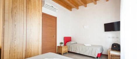 A bedroom with a wooden wardrobe, a bed with a red headboard, a bedside table with a lamp, and a TV mounted on the wall.