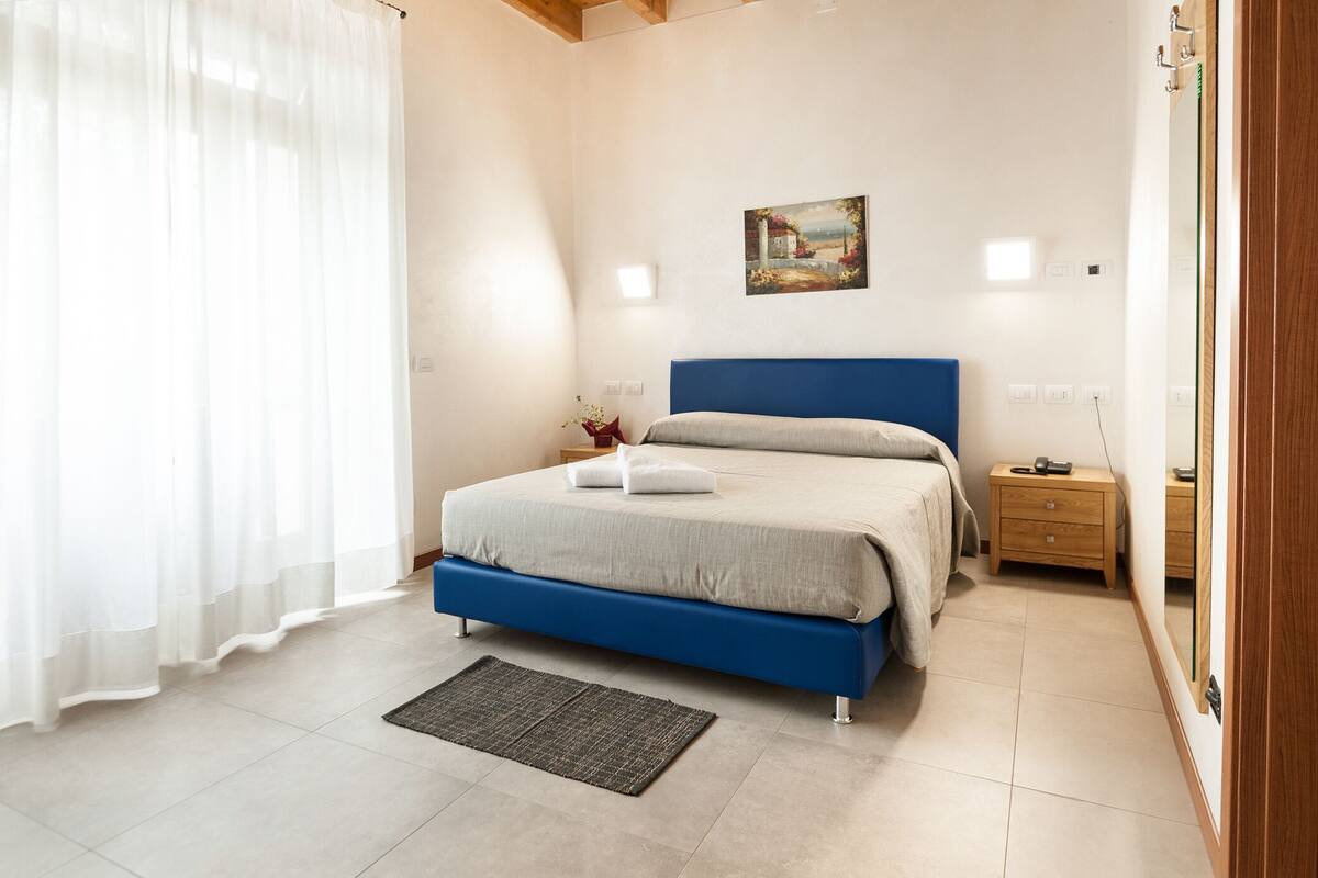 A bedroom with a bed, bedside tables, a nightstand, a painting on the wall, and a window with curtains.