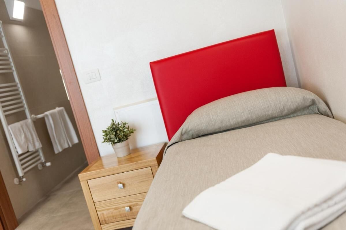 A hotel room with a bed, a nightstand, a red headboard, a small plant, and a towel.