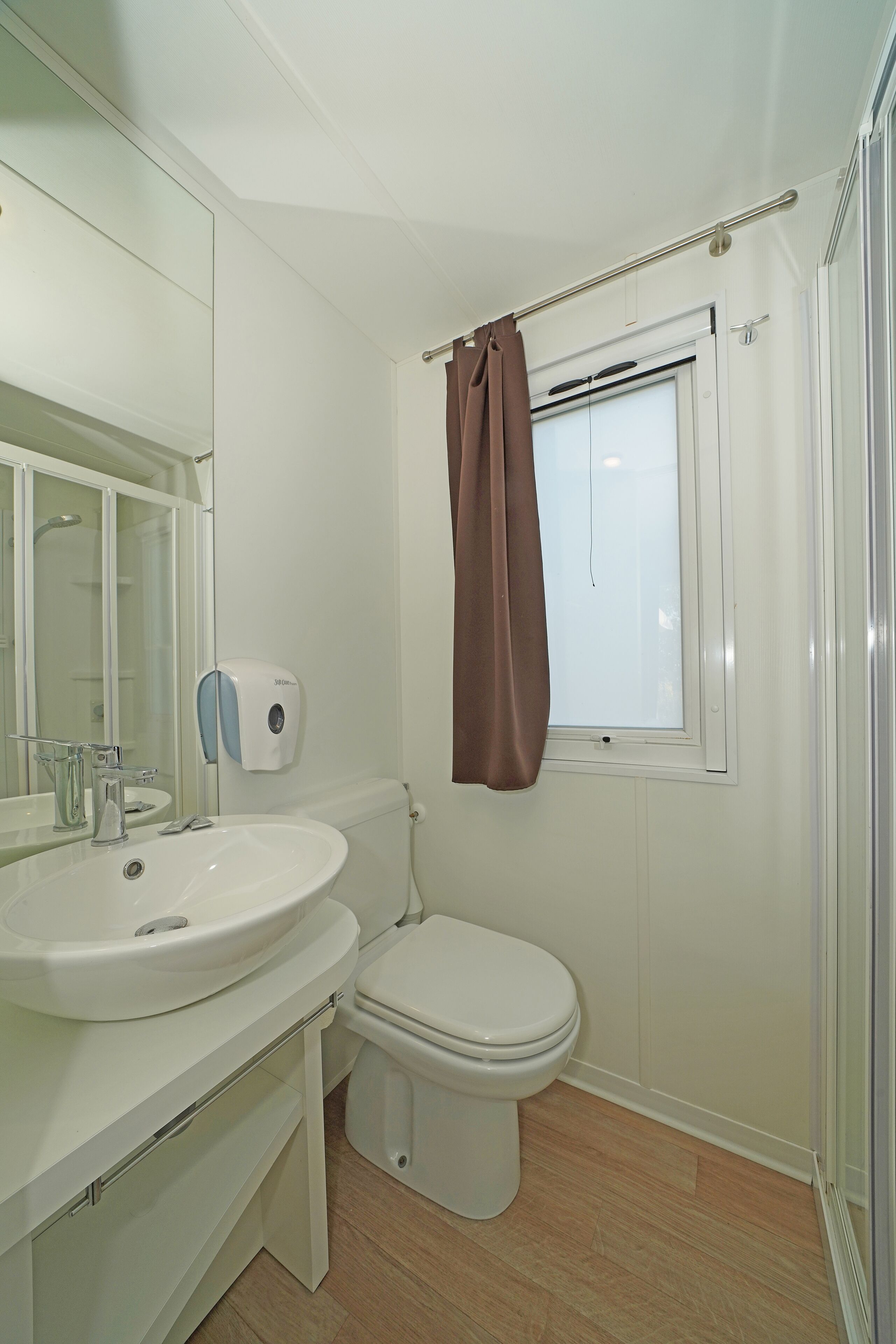 A compact bathroom with a white sink, toilet, and a window with a brown curtain.