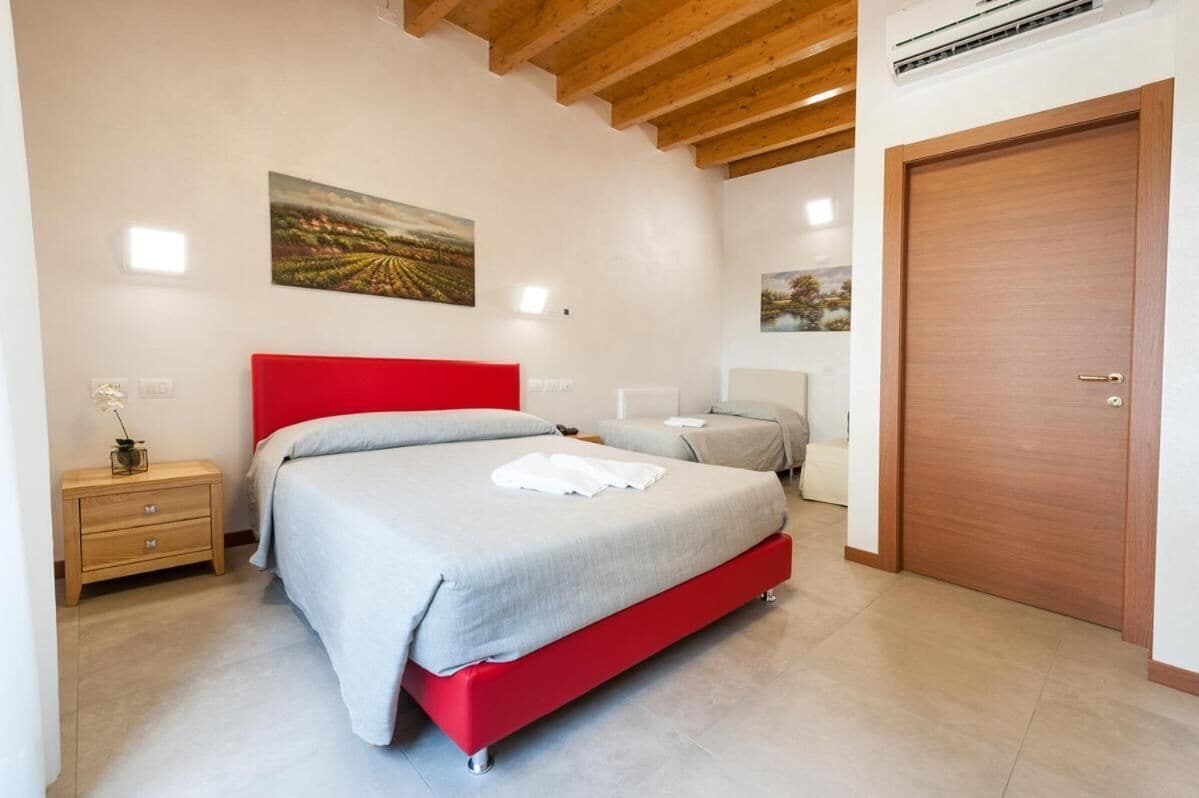 Une chambre à coucher avec un cadre de lit rouge, une couvre-lit grise, une table de chevet en bois avec un vase, et une porte avec une poignée.