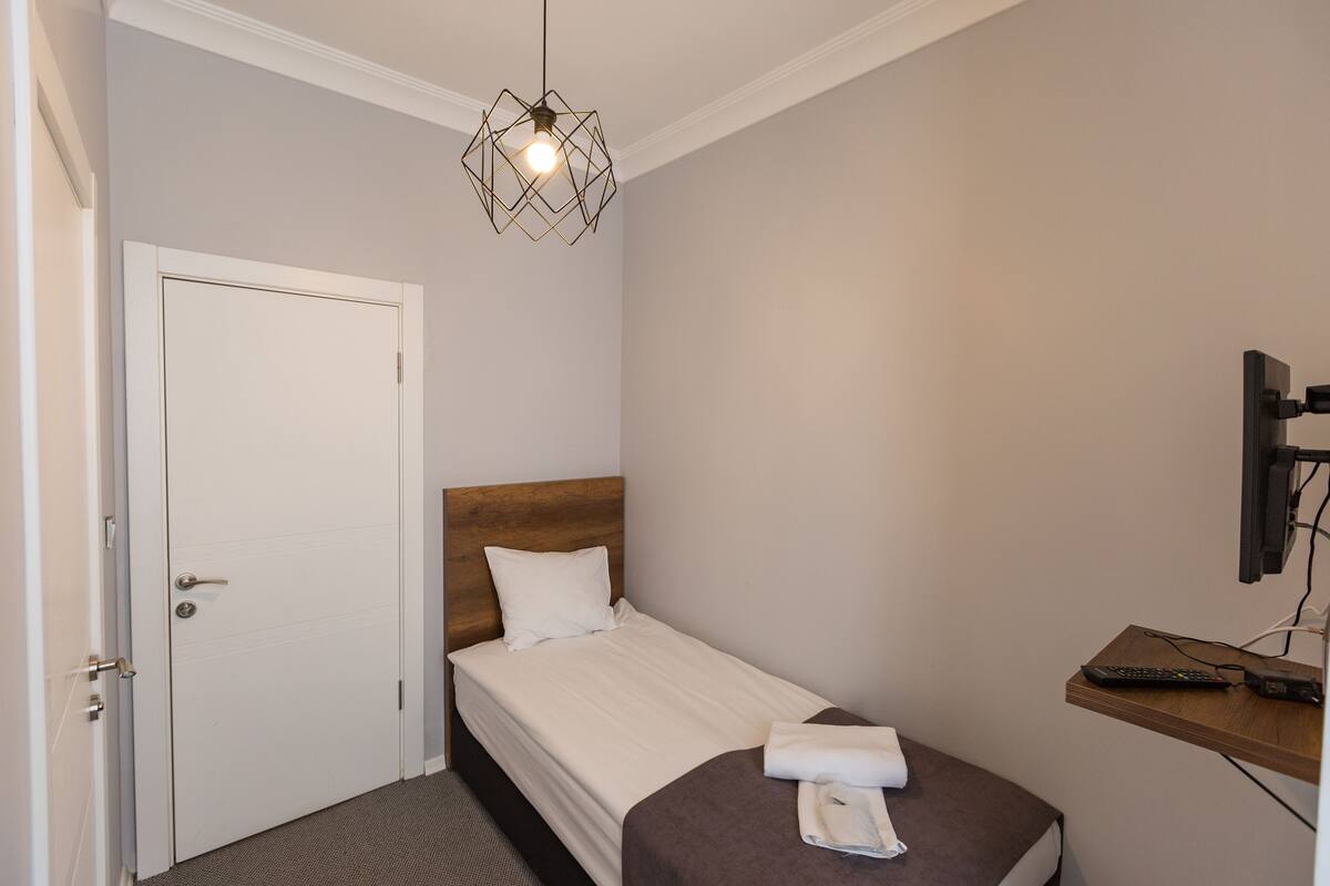 A hotel room with a single bed, a wooden headboard, a white door, a hanging light fixture, a TV mounted on the wall, and a small wooden shelf with a remote control.