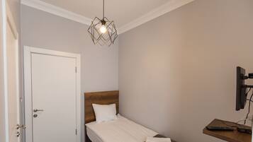 Habitación de hotel con una cama individual, cabecera de madera, puerta blanca, lámpara colgante, televisor montado en la pared y un pequeño estante con un control remoto.
