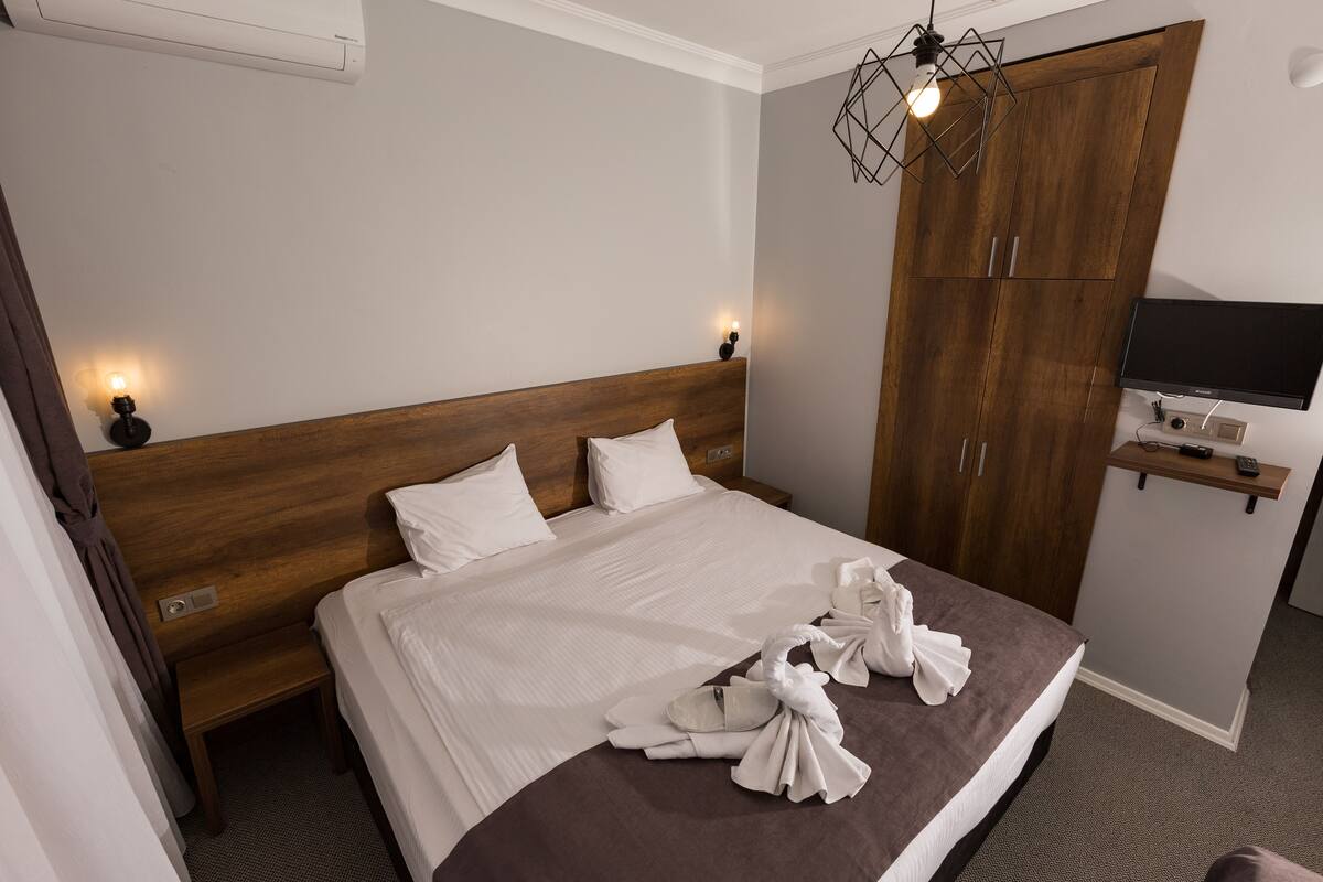 A neatly made bed with white linens, a wooden headboard, and a brown accent at the foot. There are two bedside lamps, a TV mounted on the wall, and a wooden wardrobe.
