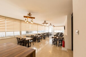 Un comedor amplio con varias mesas y sillas, ventanas grandes con persianas e iluminación colgante.