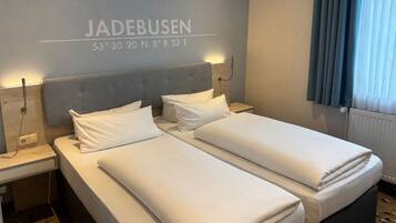 Camera d'albergo con due letti, una testiera grigia, biancheria da letto bianca, una lampada a parete e una finestra con tende.