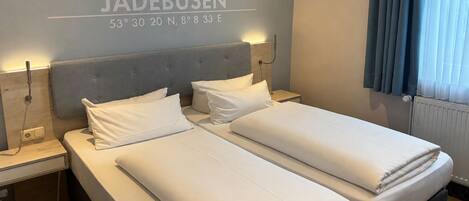 A hotel room with two beds, a grey headboard, white bedding, a wall-mounted light, and a window with curtains.