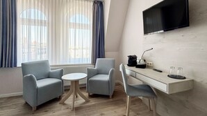 Habitación de hotel moderna con televisor de pantalla plana montado en la pared, una mesita redonda pequeña, dos sillones, un escritorio con cafetera y vistas a la ciudad a través de amplias ventanas con cortinas translúcidas.