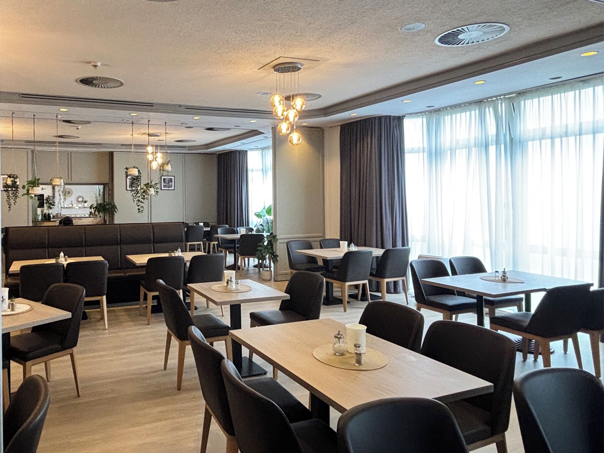 Ein moderner Essbereich mit mehreren Tischen und Stühlen, eine Bar und große Fenster mit Vorhängen.