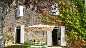 Un espace extérieur aménagé pour les repas, avec une table et des chaises, un grand parasol et, en arrière-plan, un bâtiment en pierre.