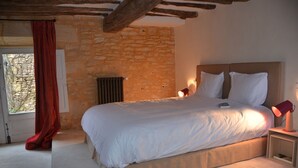 Une chambre avec un mur en pierre, un lit recouvert de draps blancs, une lampe rouge et une fenêtre ornée d’un rideau rouge.