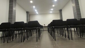 Ein Konferenzraum mit Reihen schwarzer Stühle, einem an der Decke befestigten Projektor und einem gefliesten Boden.