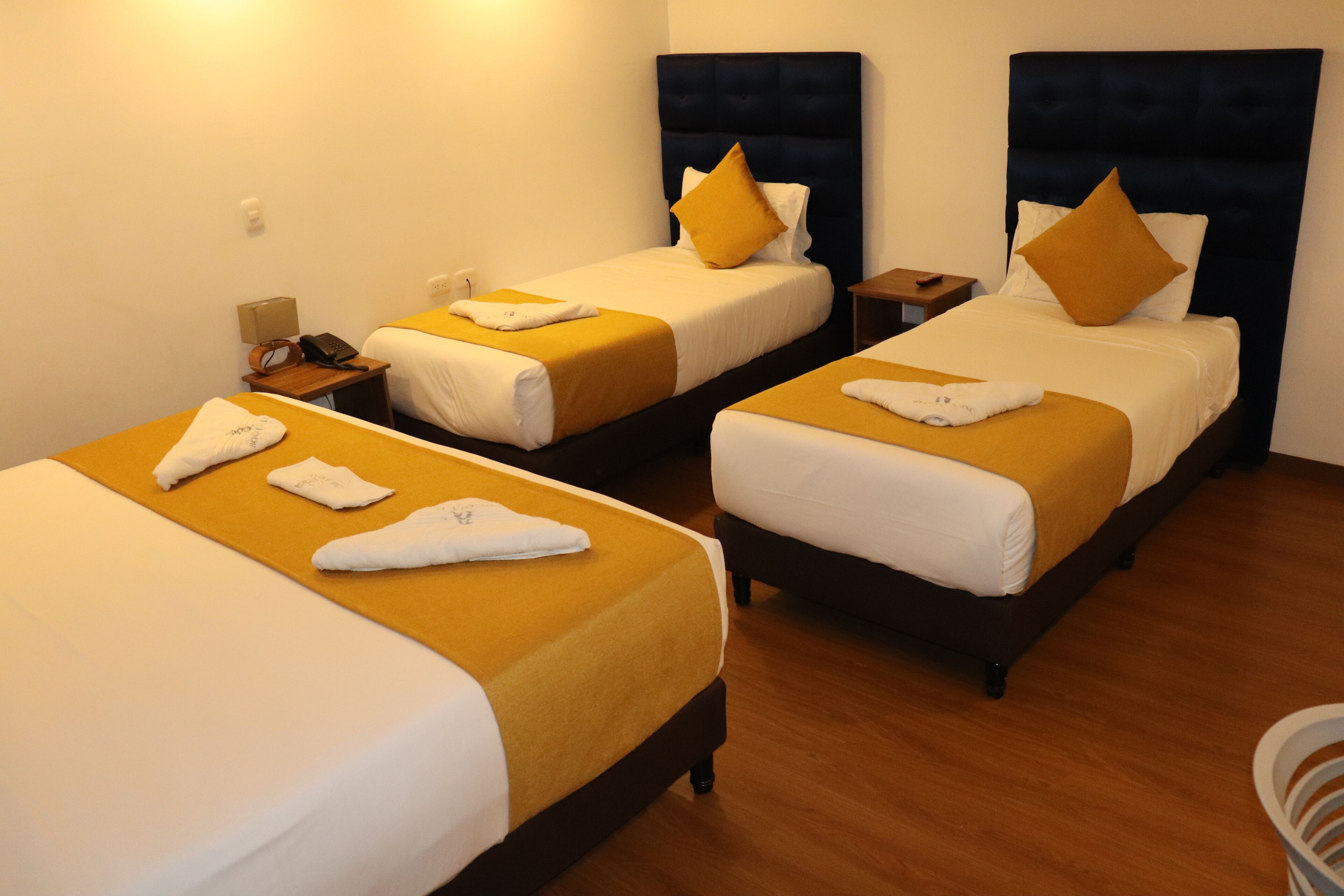 Quarto de hotel com duas camas de solteiro, cada uma com colcha amarela e almofadas brancas, e uma mesa de cabeceira de madeira com luminária e telefone.