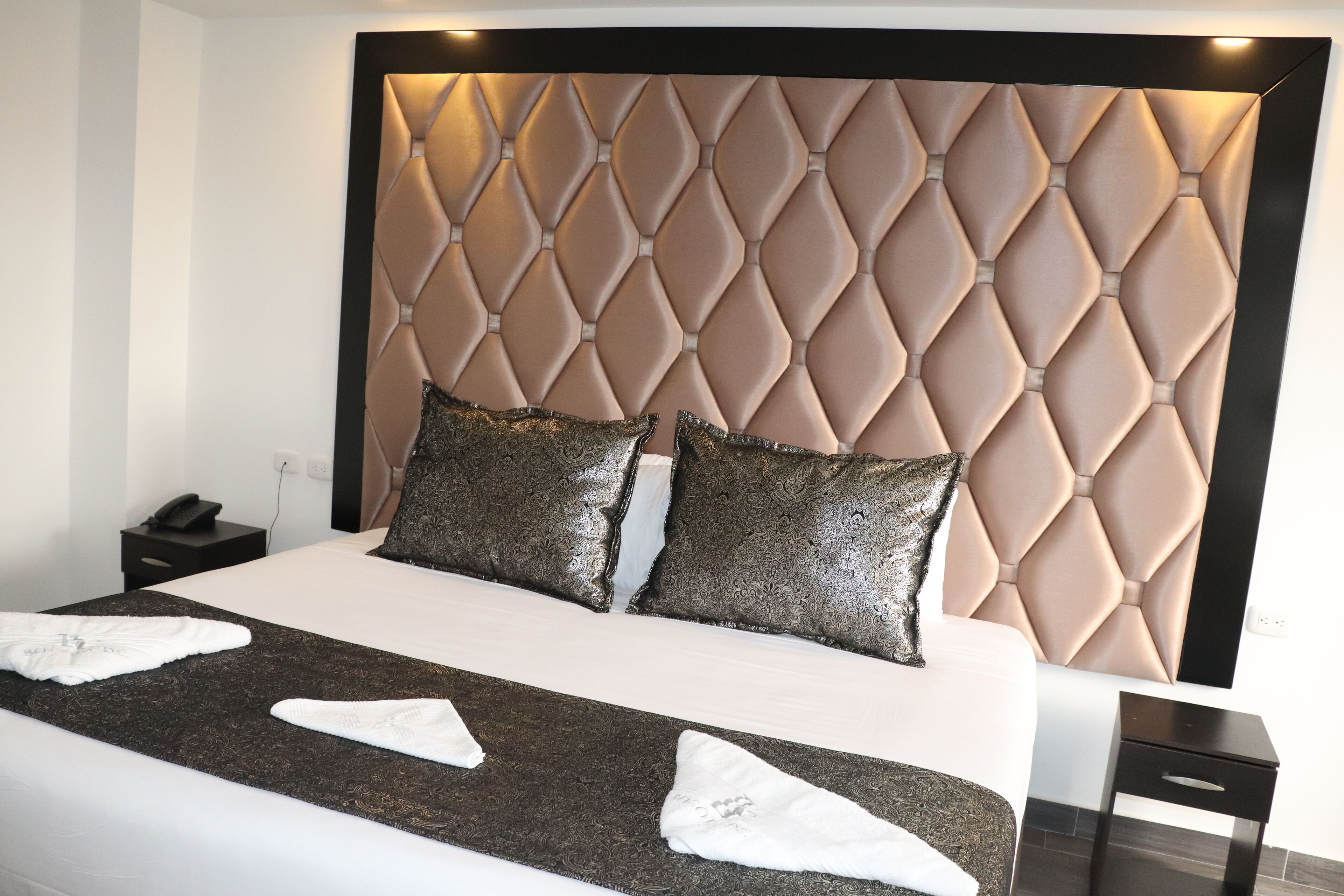 Habitación de hotel con una cama grande, cabecera con diseño de rombos, dos almohadas decorativas y una mesita de noche con un teléfono.