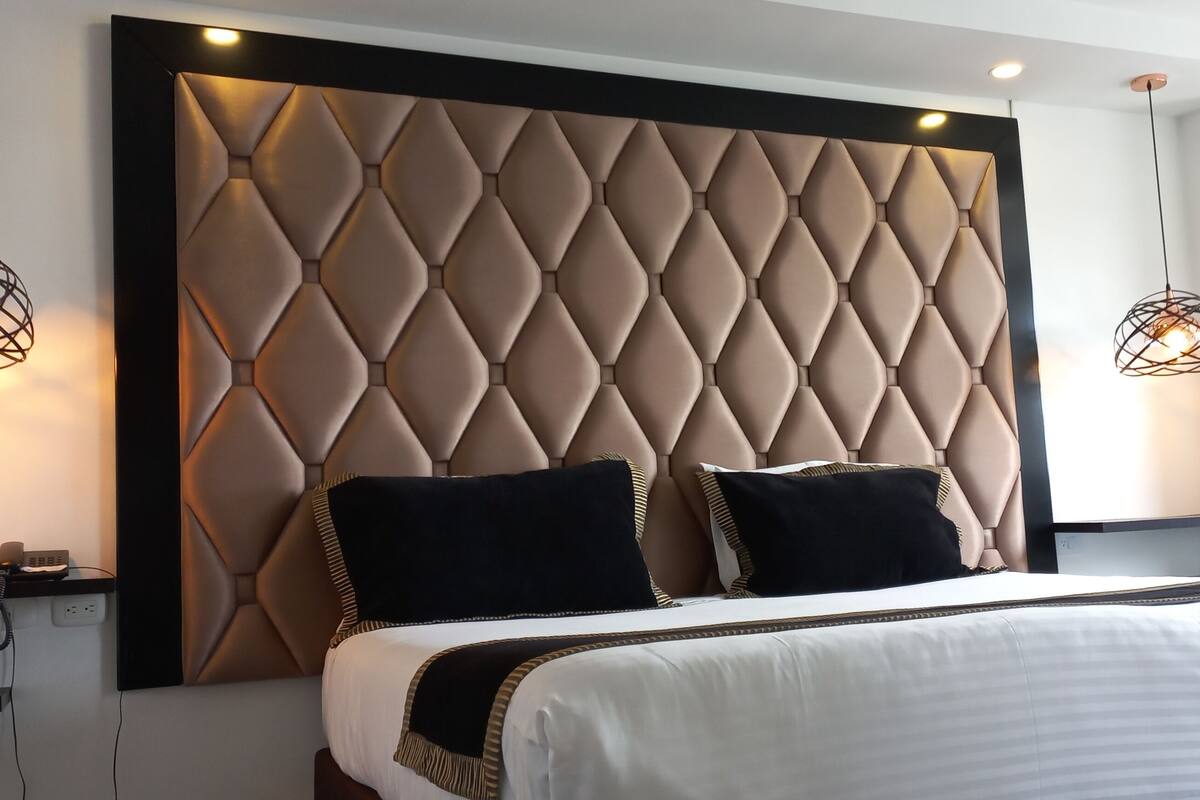 A modern hotel room with a tufted headboard, a large bed with white bedding, two black pillows, and a black throw blanket. There are two pendant lights with a geometric design on either side of the bed.