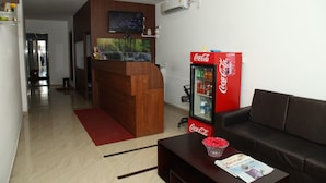A modern living area with a wooden reception desk, a large flat-screen TV, a Coca-Cola refrigerator, a leather sofa, and a coffee table with a magazine. - Sai Arya Lodging & Hall (Mangaluru)