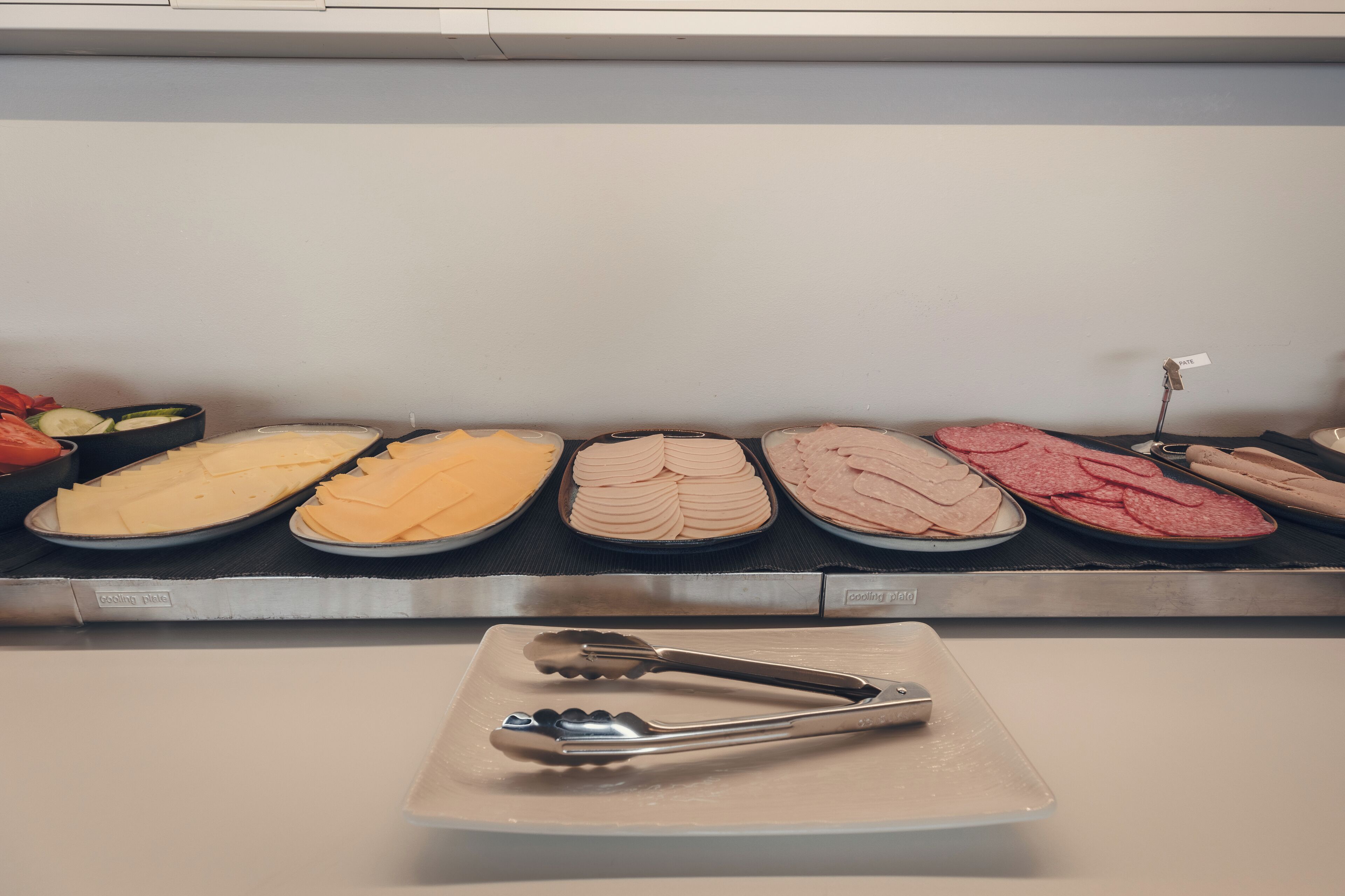 A display of sliced cheeses and meats on trays, with tongs placed on a plate in the foreground.