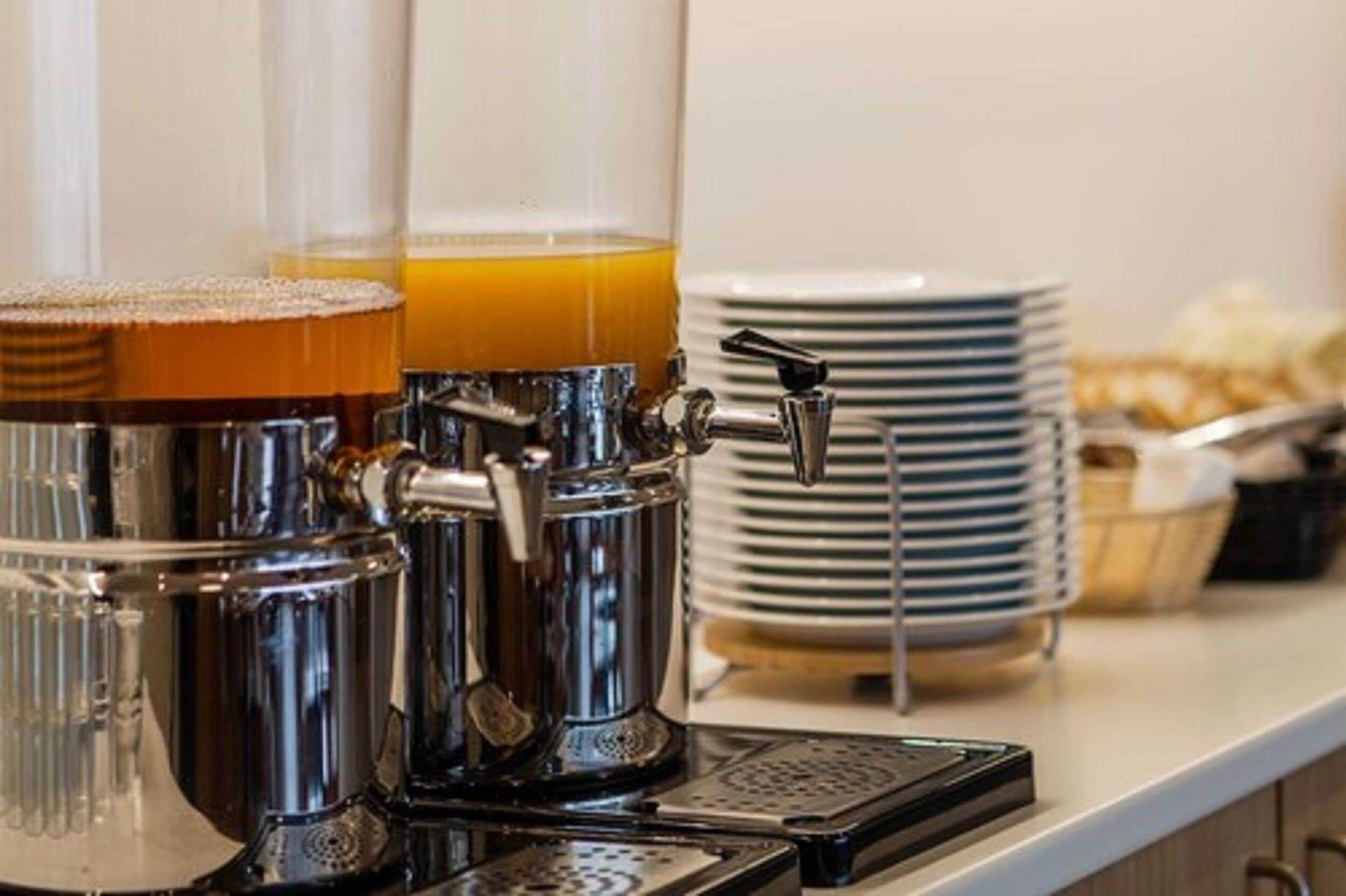 A coffee machine with multiple dispensers, a stack of plates, and a basket of pastries on a counter.