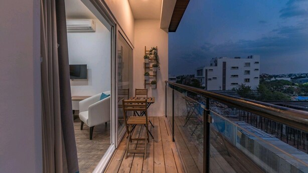 Un balcón con piso de madera, una mesita y sillas. Cuenta con baranda y vistas a edificios en la distancia.