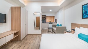 Una habitación de hotel moderna con cama, mesita de noche, un escritorio con silla, espejo, armario y una pequeña cocina en el fondo.