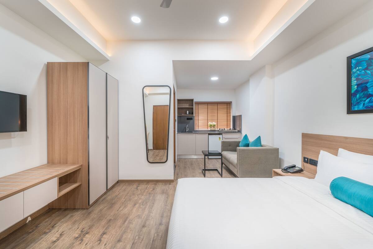Quarto de hotel moderno com cama, mesa de cabeceira, escrivaninha com cadeira, espelho, guarda-roupa e uma kitchenette ao fundo.