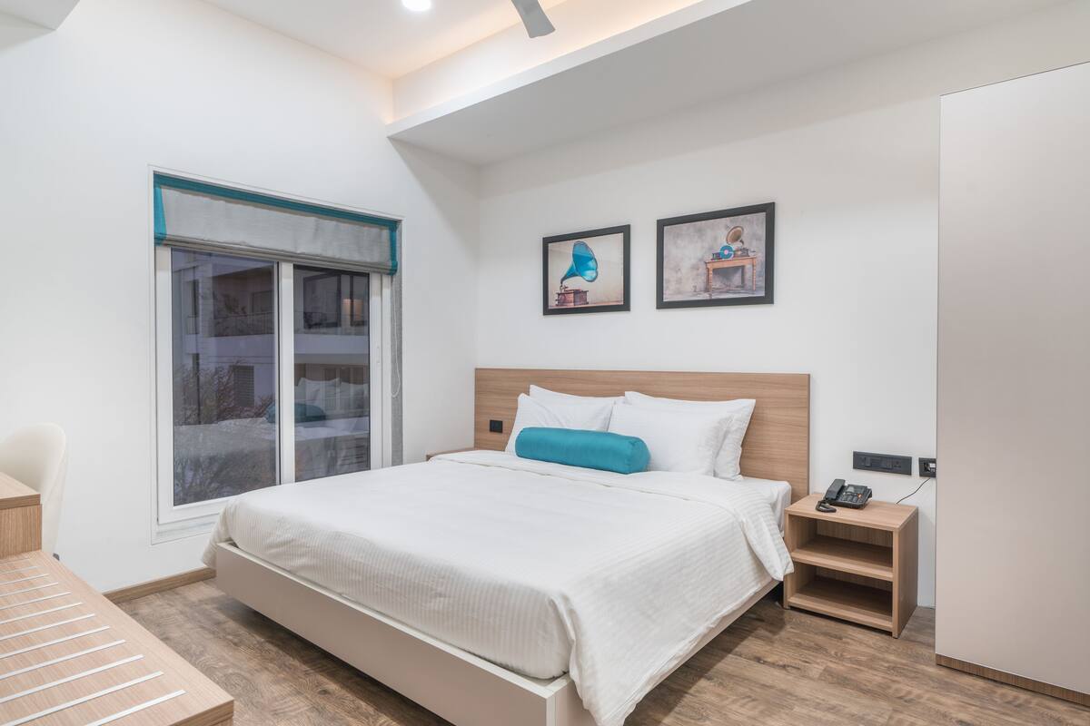 Quarto moderno com cama grande, mesa de cabeceira com telefone e janela com persianas.
