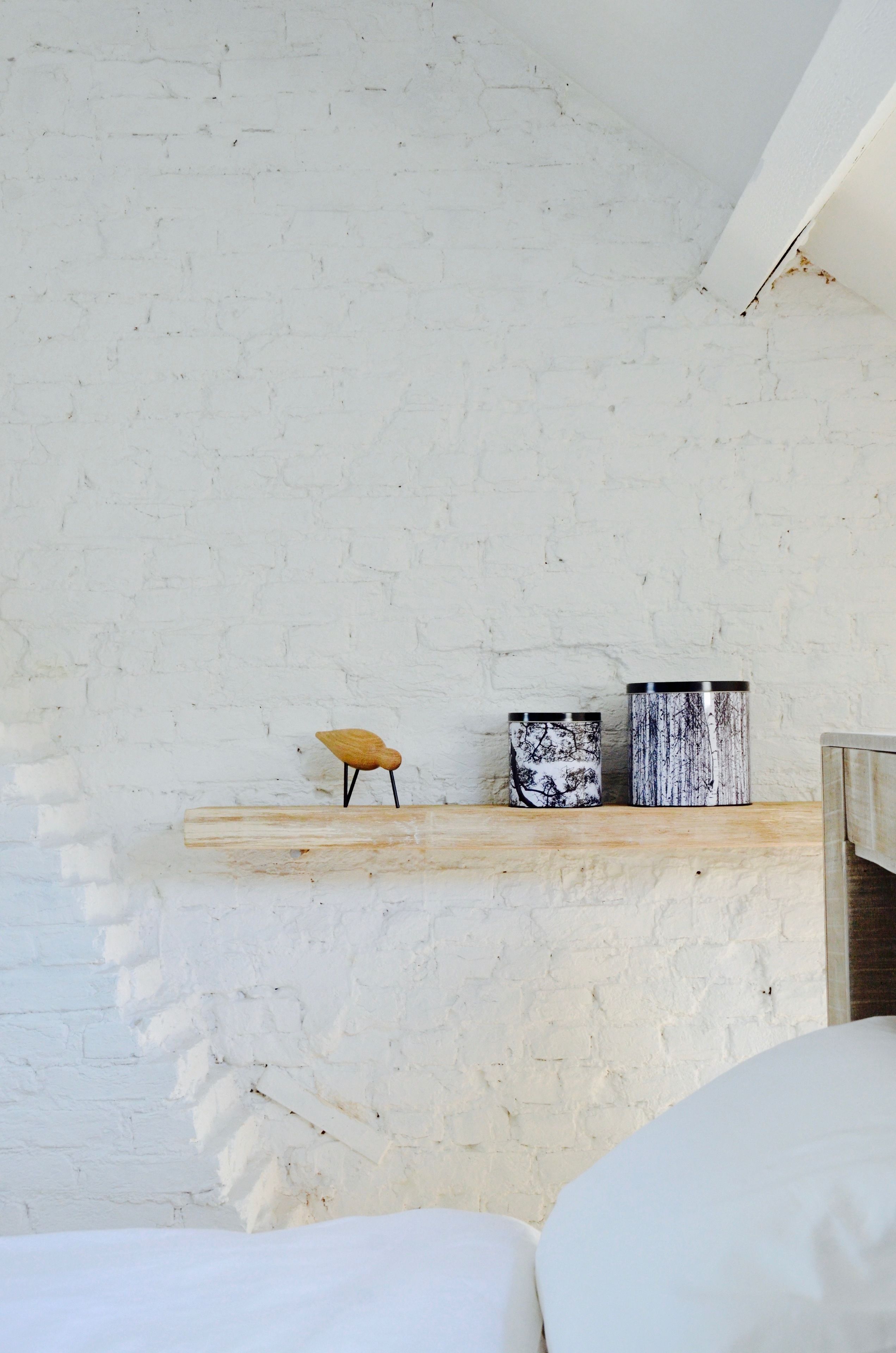 A white brick wall with a wooden shelf holding a small wooden stool and two cylindrical containers.