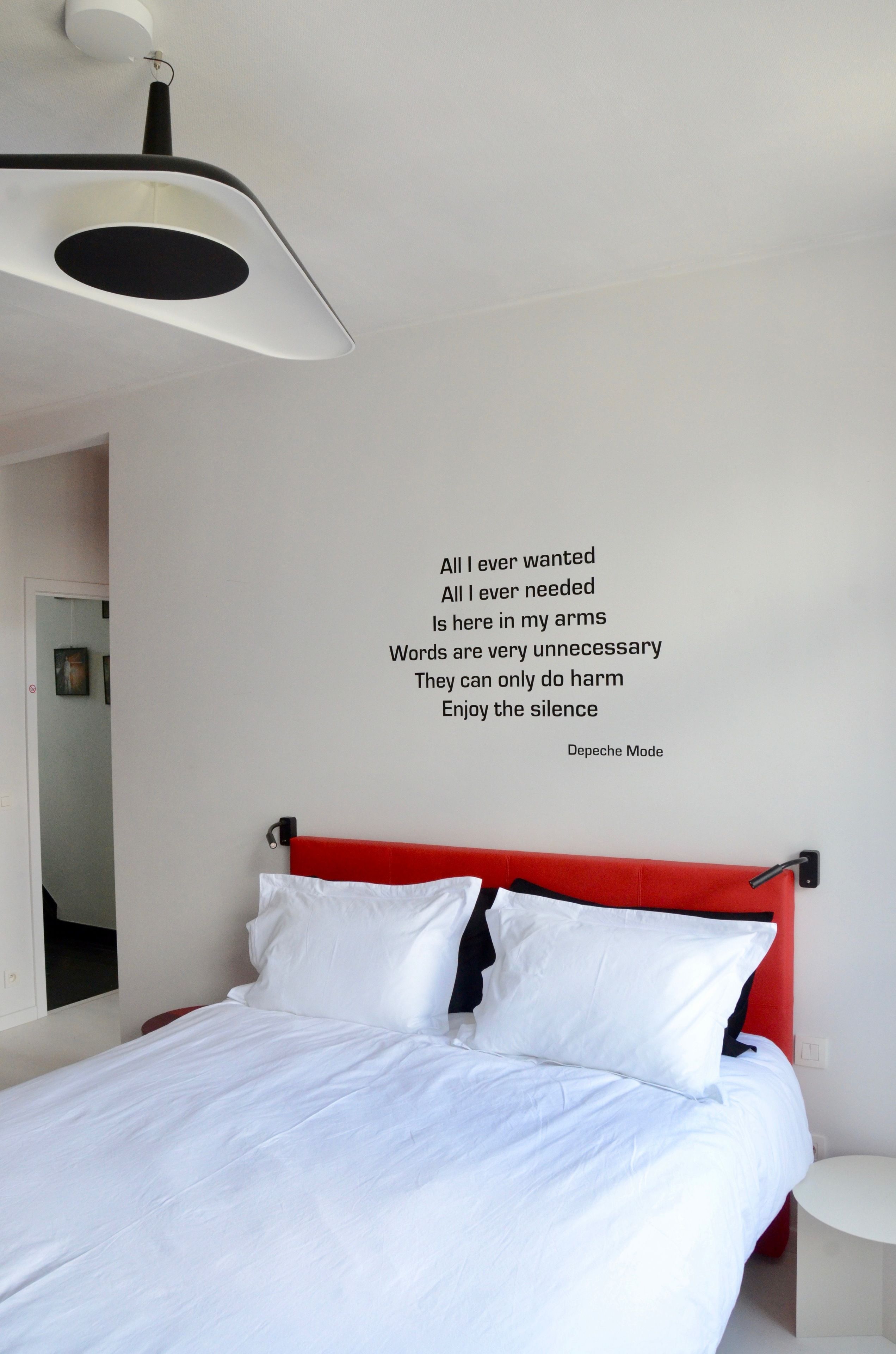 Ein modernes Schlafzimmer mit einem weißen Bett, einer roten Kopfteil und einem an der Wand befestigten Zitat von Depeche Mode.