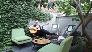 Un homme joue de la guitare sur une terrasse où se trouvent des chaises vertes et une petite table.