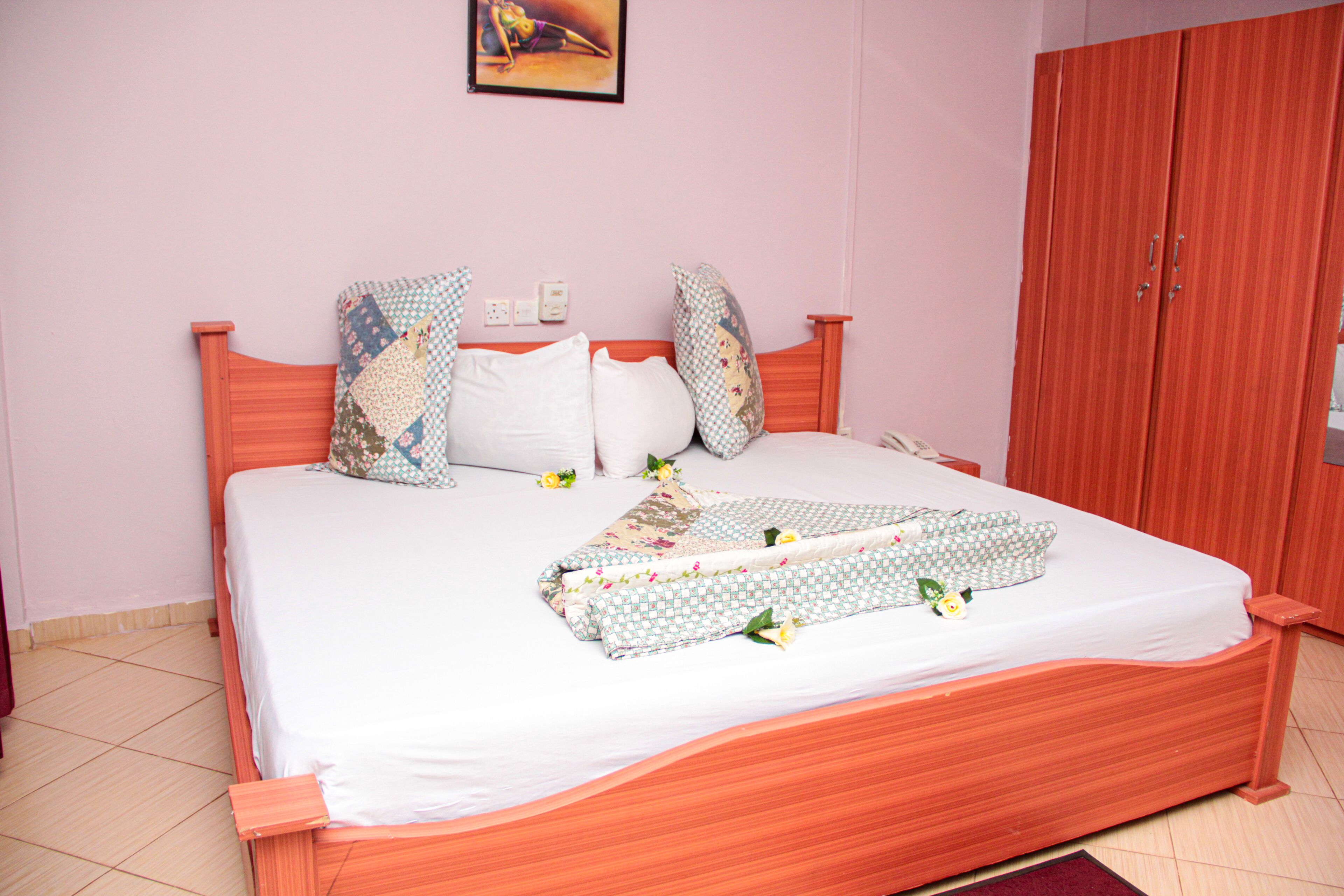 A neatly made bed with a patterned bedspread and pillows, a wooden headboard, and a red wardrobe in a room with pink walls.