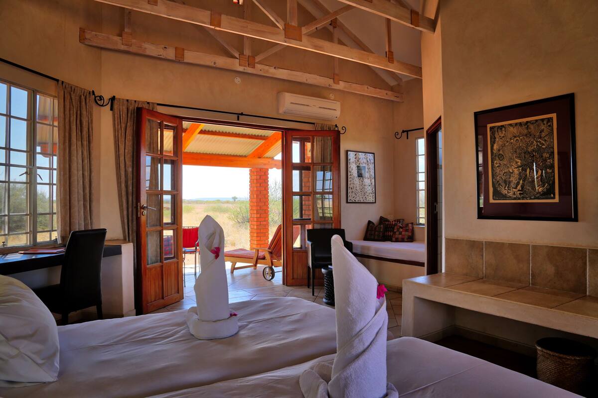 Habitación de hotel con dos camas, un escritorio, una silla y un banco. Se ve una puerta que da a un espacio al aire libre con vistas al paisaje.