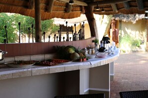 An outdoor bar area with a thatched roof, a variety of fruits and condiments on the counter, and a rustic setting. - Guest Farm Kiripotib (Windhoek)