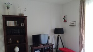 A living room with a TV on a stand, a bookshelf, a floor lamp, and a small table with a red cushion. - T2 curists classified 3 * Res secure 10 minutes walk from the thermal baths (Lamalou-les-Bains)