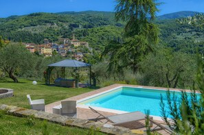 A swimming pool with lounge chairs, a gazebo, and a view of a hillside village.