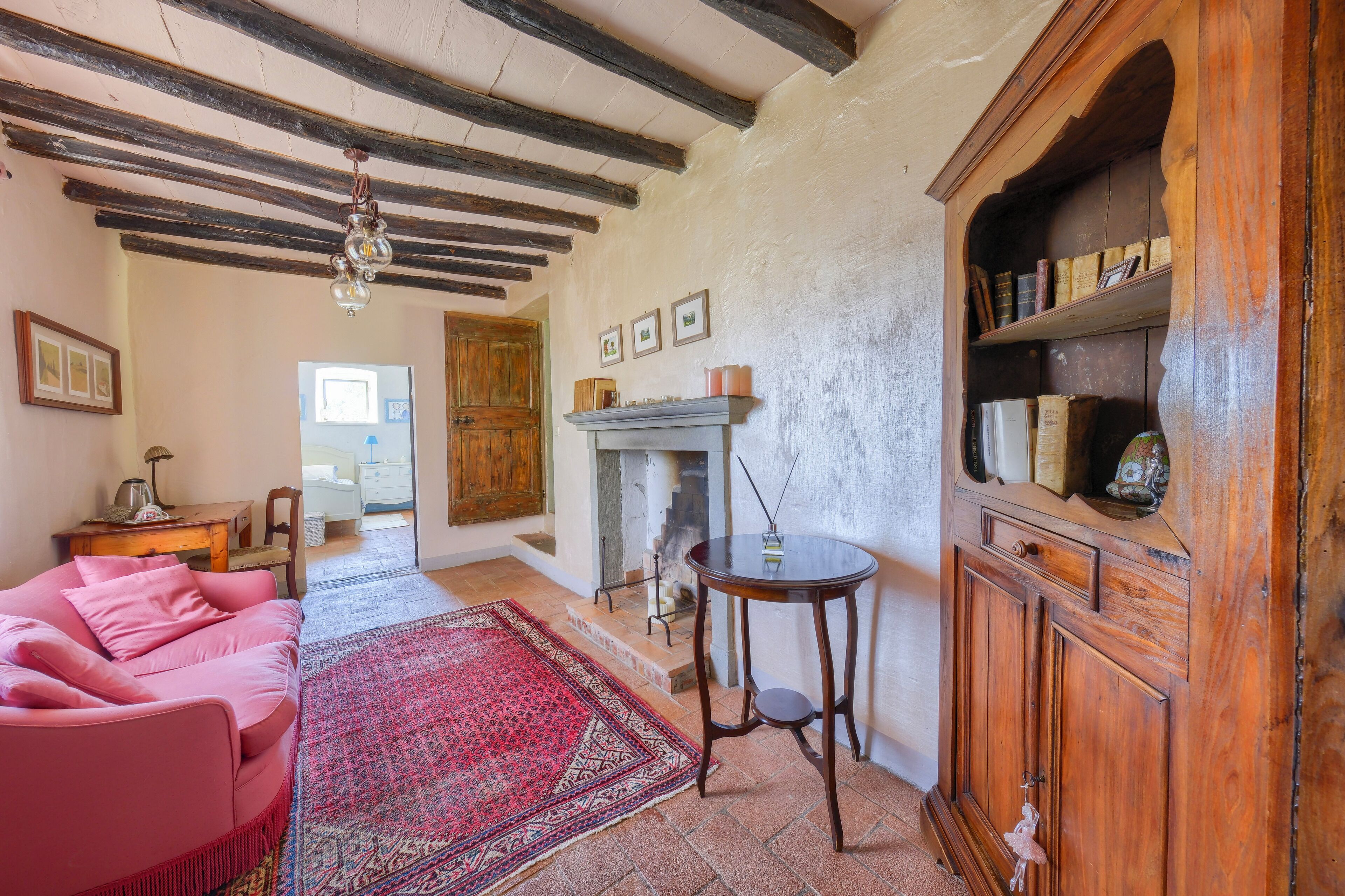 Um quarto acolhedor com teto de madeira, um sofá rosa, um tapete vermelho estampado e um armário de madeira com livros e objetos decorativos.