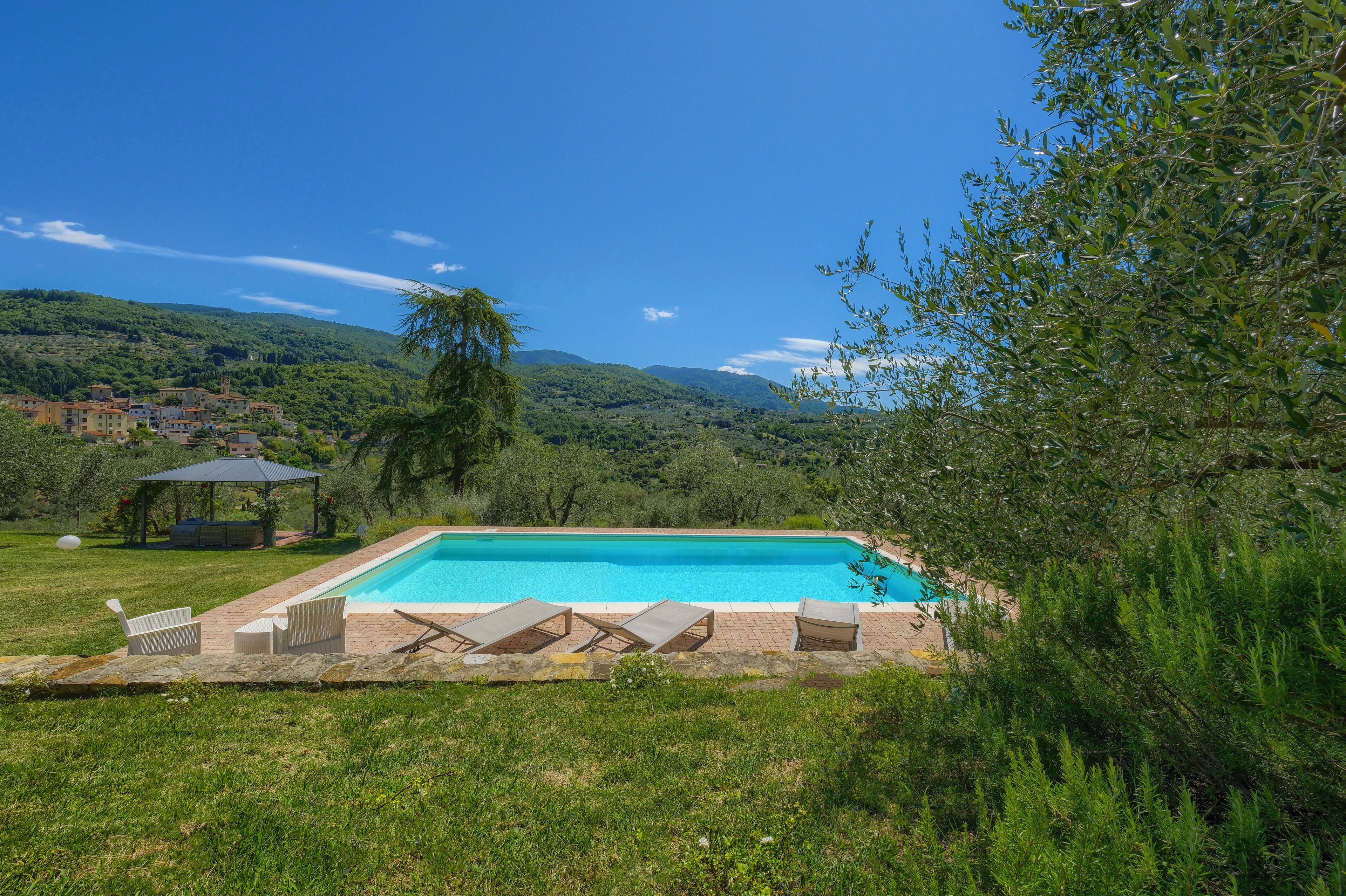 Una piscina rettangolare con poltrone a sdraio e un gazebo immersa in un paesaggio verde e rigoglioso.