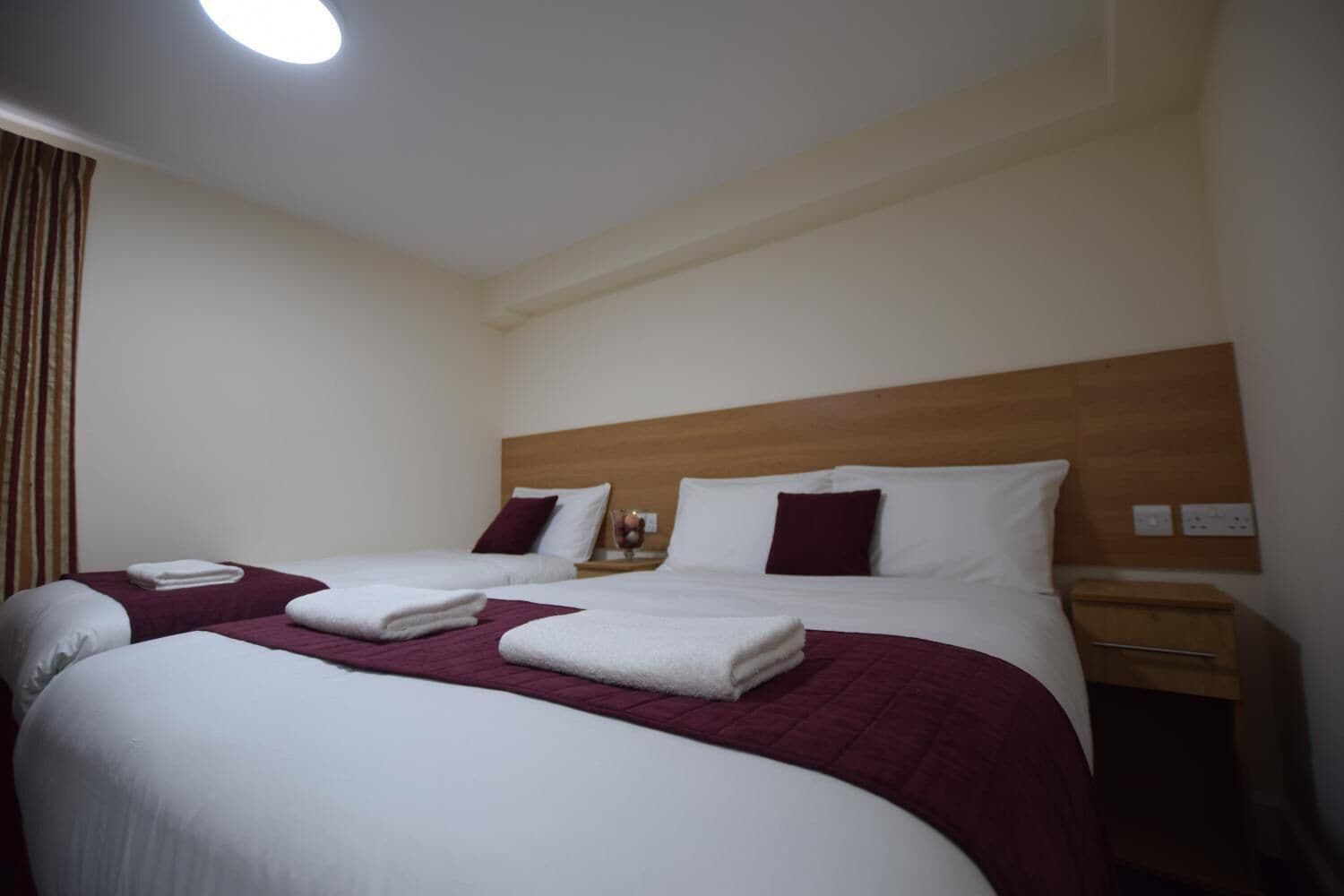 A hotel room with two beds, each with white linens and maroon blankets, a wooden headboard, and a nightstand with a lamp and a small table.