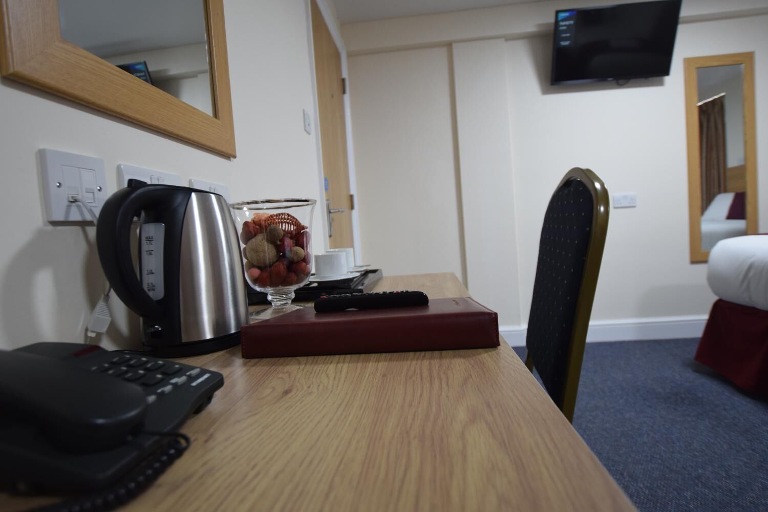 A hotel room with a wooden desk, a kettle, a bowl of fruit, a remote control, a telephone, and a bed with a red bedspread.