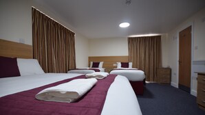 Quarto de hotel com três camas, cada uma com lençóis brancos e um cobertor vermelho. As cabeceiras e o guarda-roupa são de madeira.