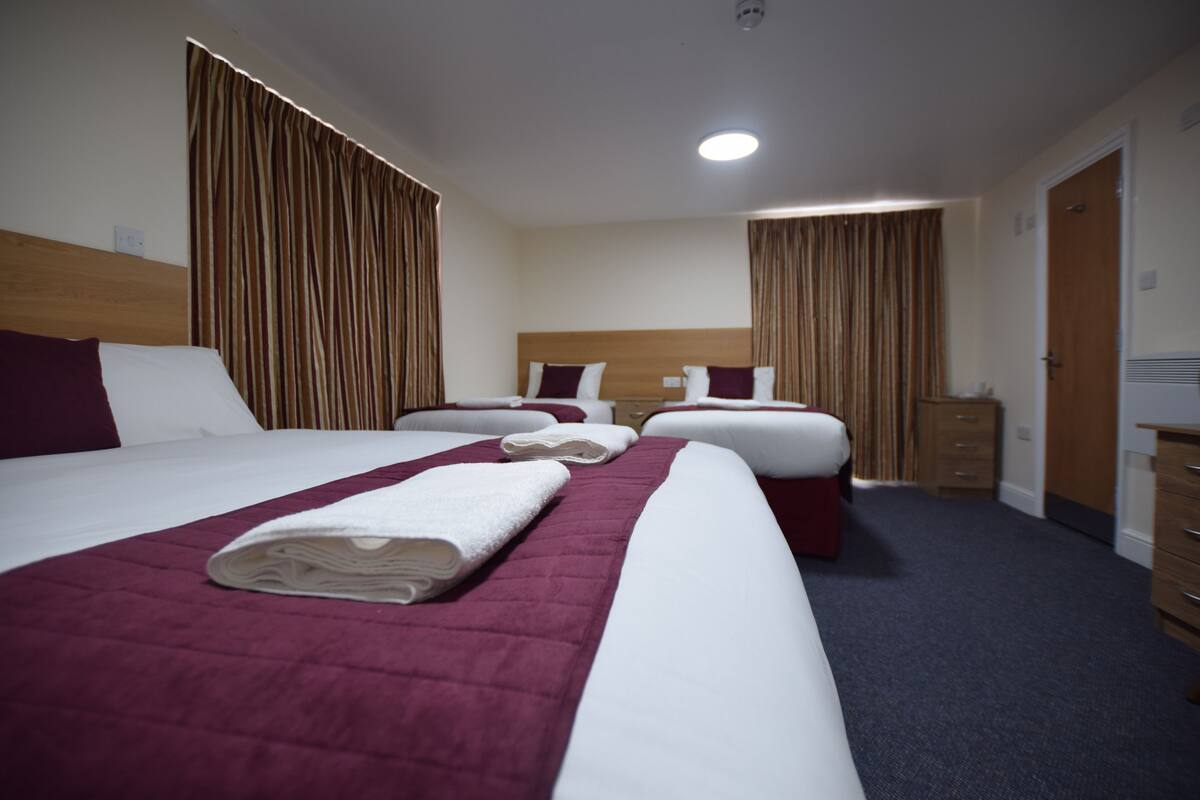 A hotel room with three beds, each with white linens and a red blanket. There are wooden headboards and a wooden wardrobe.