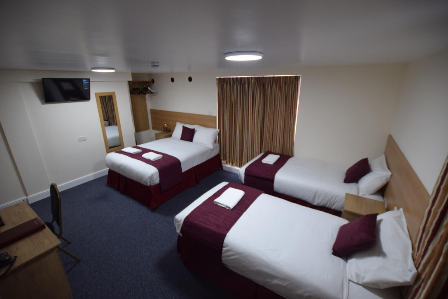 A hotel room with three single beds, a TV, a wooden headboard, and a window with curtains.